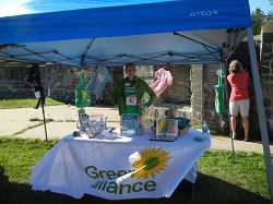 Persona standing in Green Alliance Booth(under awning)  at Zero Waste Event - tablecloth draped across front of table shows yellow daisy and the words Green Alliance
