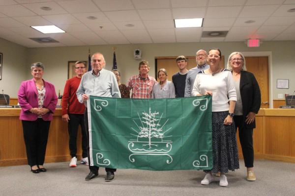 Image of the Kittery Town Council Accepting a Tribal Flag that's green with a white emblem from the Cowasuck Band of the Penacook-Abenaki People